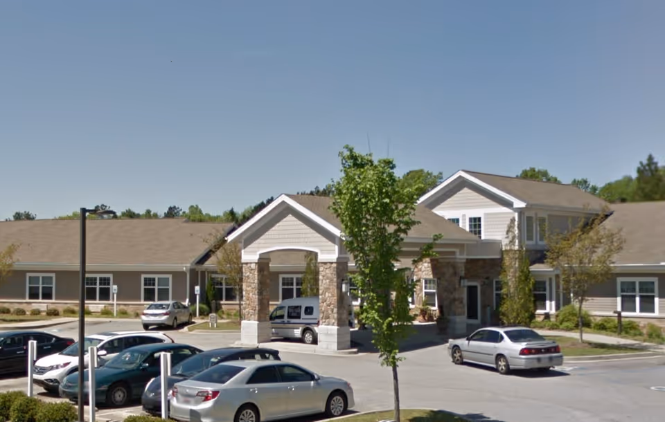 Front exterior of a single-story post-acute care building with a covered entrance, landscaping and a parking lot with several cars.