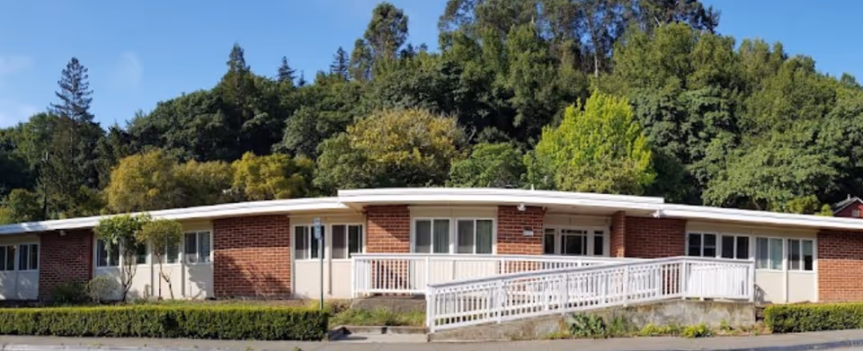 Single-story brick assisted living building with a white railing ramp and surrounding trees.