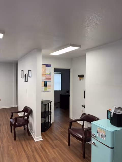 White-walled hallway in a senior living facility with chairs and a mini-fridge.
