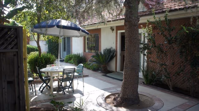 Outdoor courtyard with a table, chairs, umbrella, and a tree beside a brick building.
