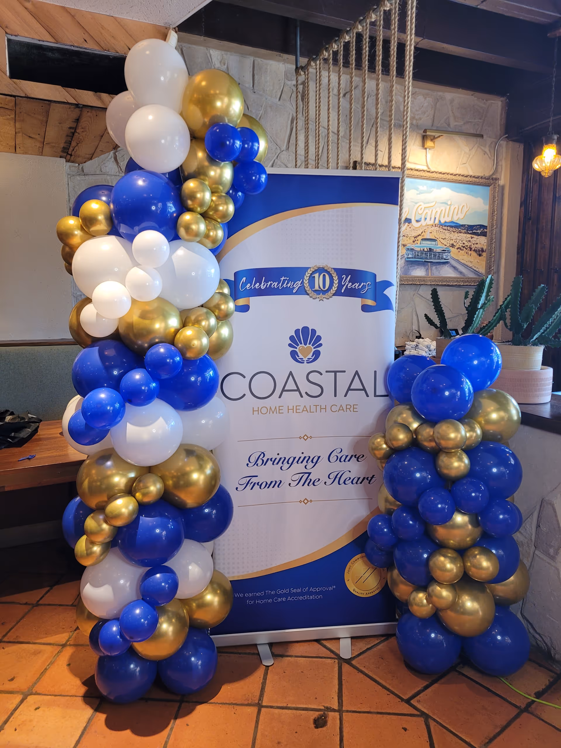 A banner for Coastal Home Health Care is flanked by blue, white, and gold balloons indoors.