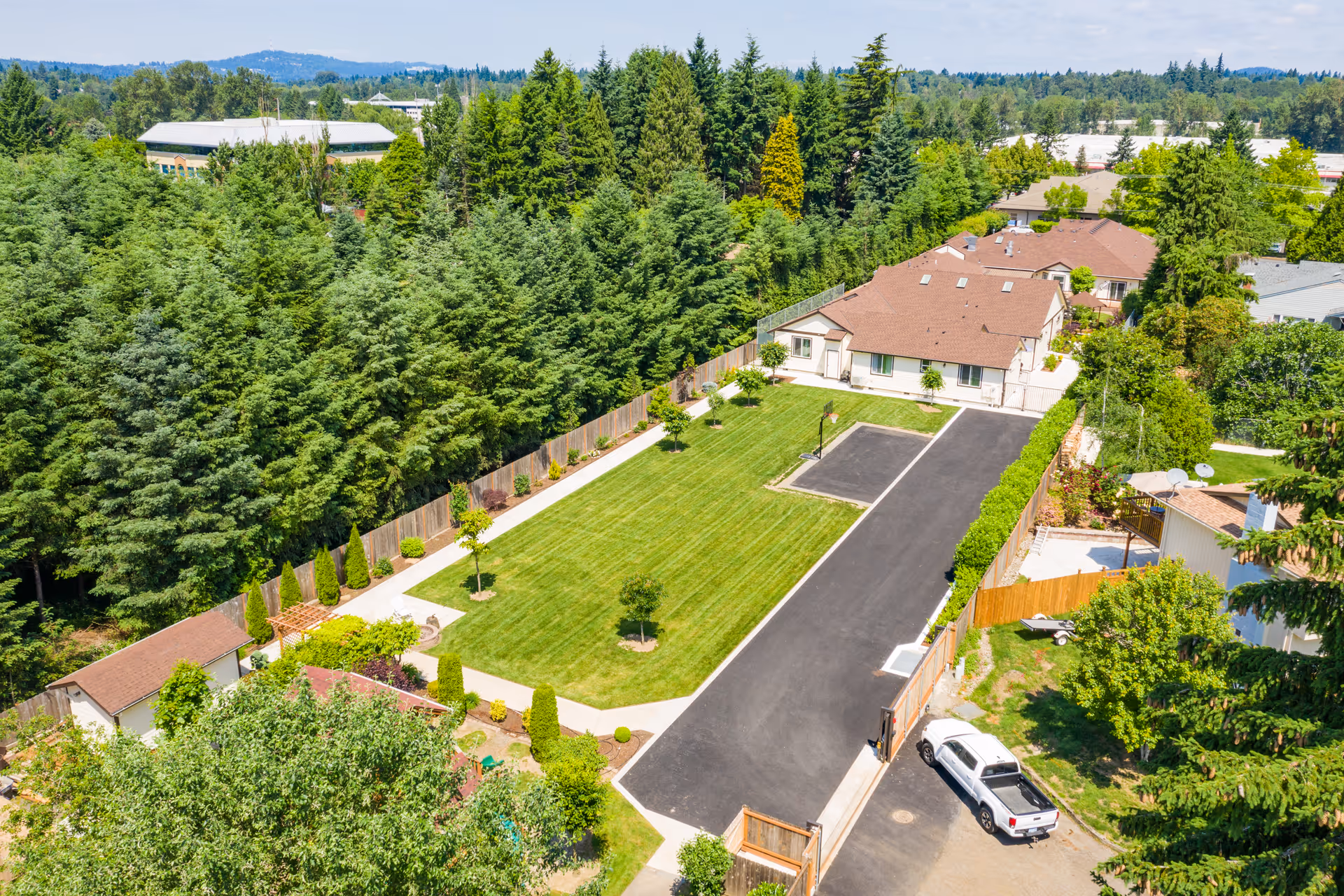 Aerial view of a senior living facility with a large green lawn, paved driveway, and several single-story buildings surrounded by trees and greenery under a clear sky.