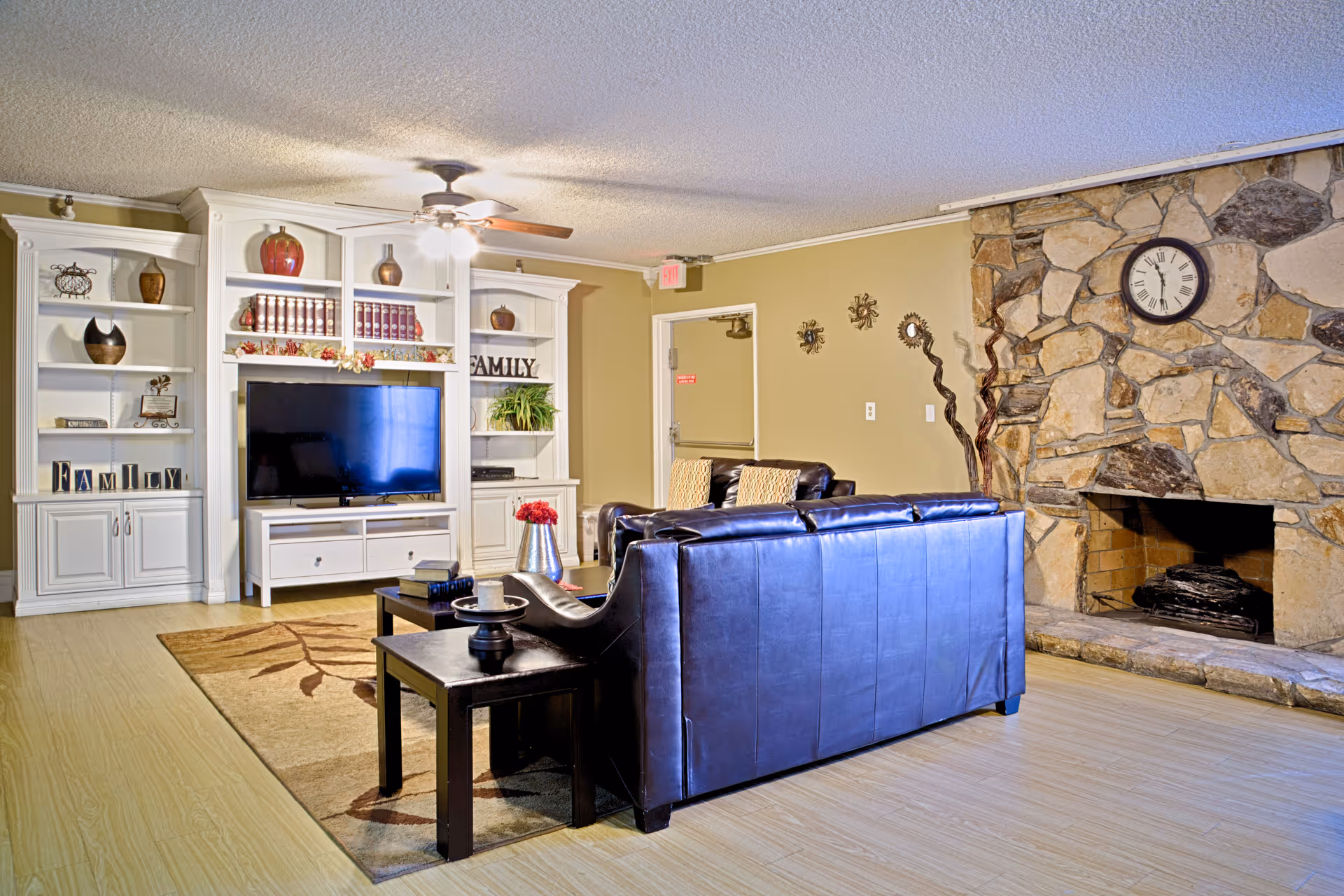 Communal living room with leather sofas facing a TV set in white built-in shelving and a stone fireplace with a clock.