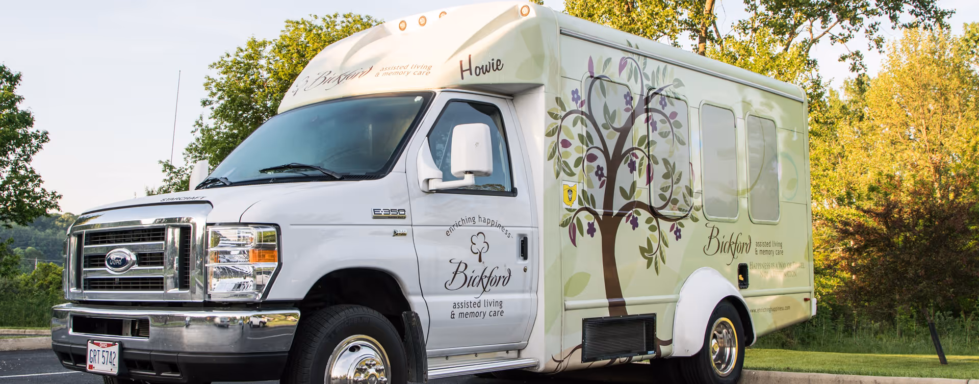 A Bickford of Iowa City branded shuttle van with a decorative tree graphic parked outdoors near trees.