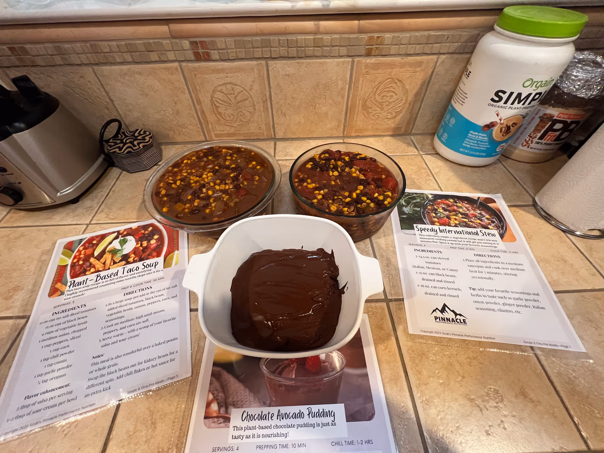Bowls of chocolate dessert on kitchen counter with colorful toppings beside recipe sheets.