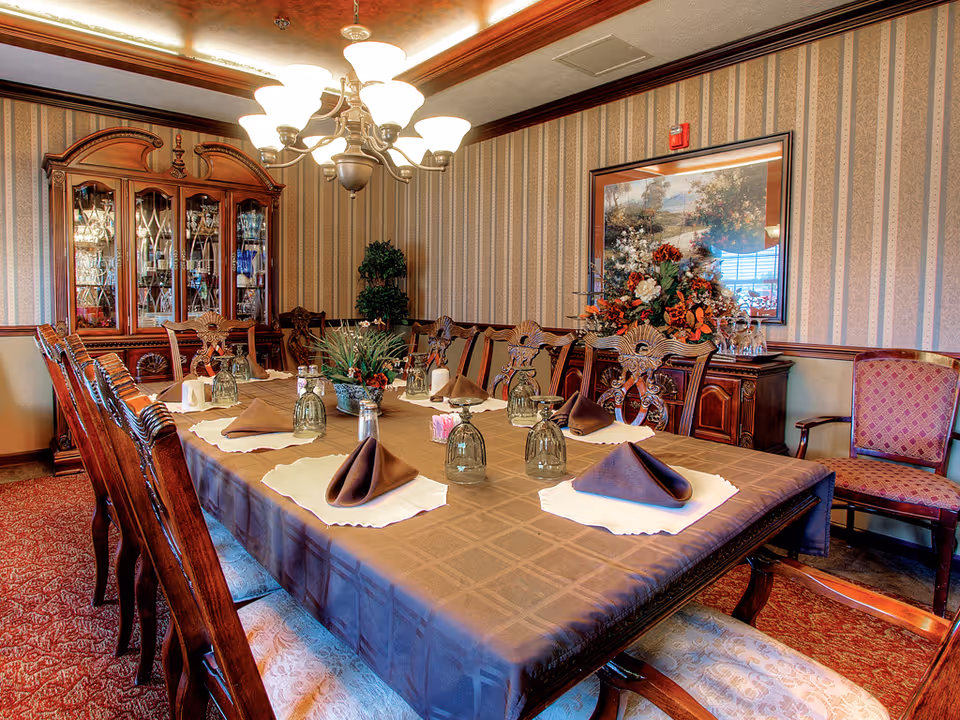 A formal dining room with a long wooden table covered with a brown tablecloth and set with inverted glasses and folded brown napkins on white placemats. The room features ornate wooden chairs, a chandelier overhead, a glass-fronted china cabinet filled with glassware, a sideboard with a floral arrangement, and patterned wallpaper.