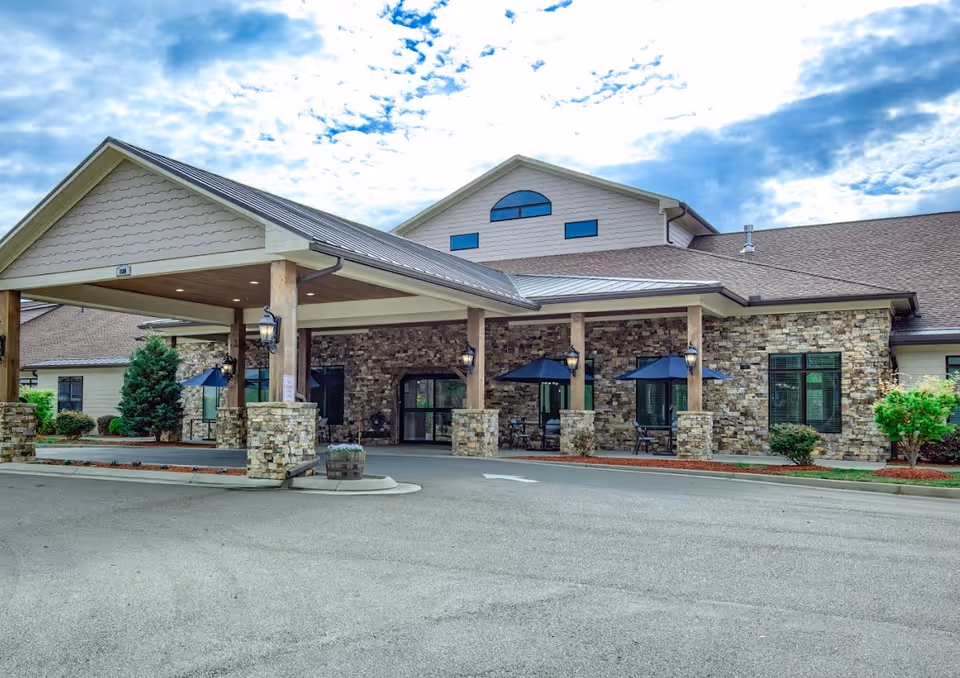Exterior view of Blue Ridge Assisted Living and Memory Care building featuring a covered entrance with stone pillars, outdoor seating with umbrellas, and a partly cloudy sky.