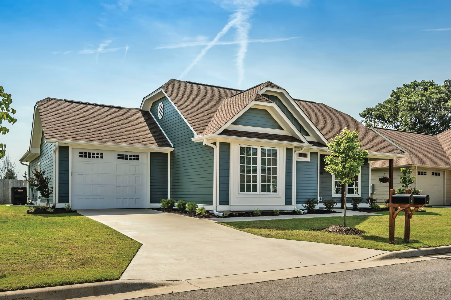 Exterior view of a single-story residential building with blue siding and a brown shingled roof. The house features a garage with a white door, a concrete driveway, a small front yard with green grass, a young tree, and a wooden mailbox stand. The sky is clear with some wispy clouds.
