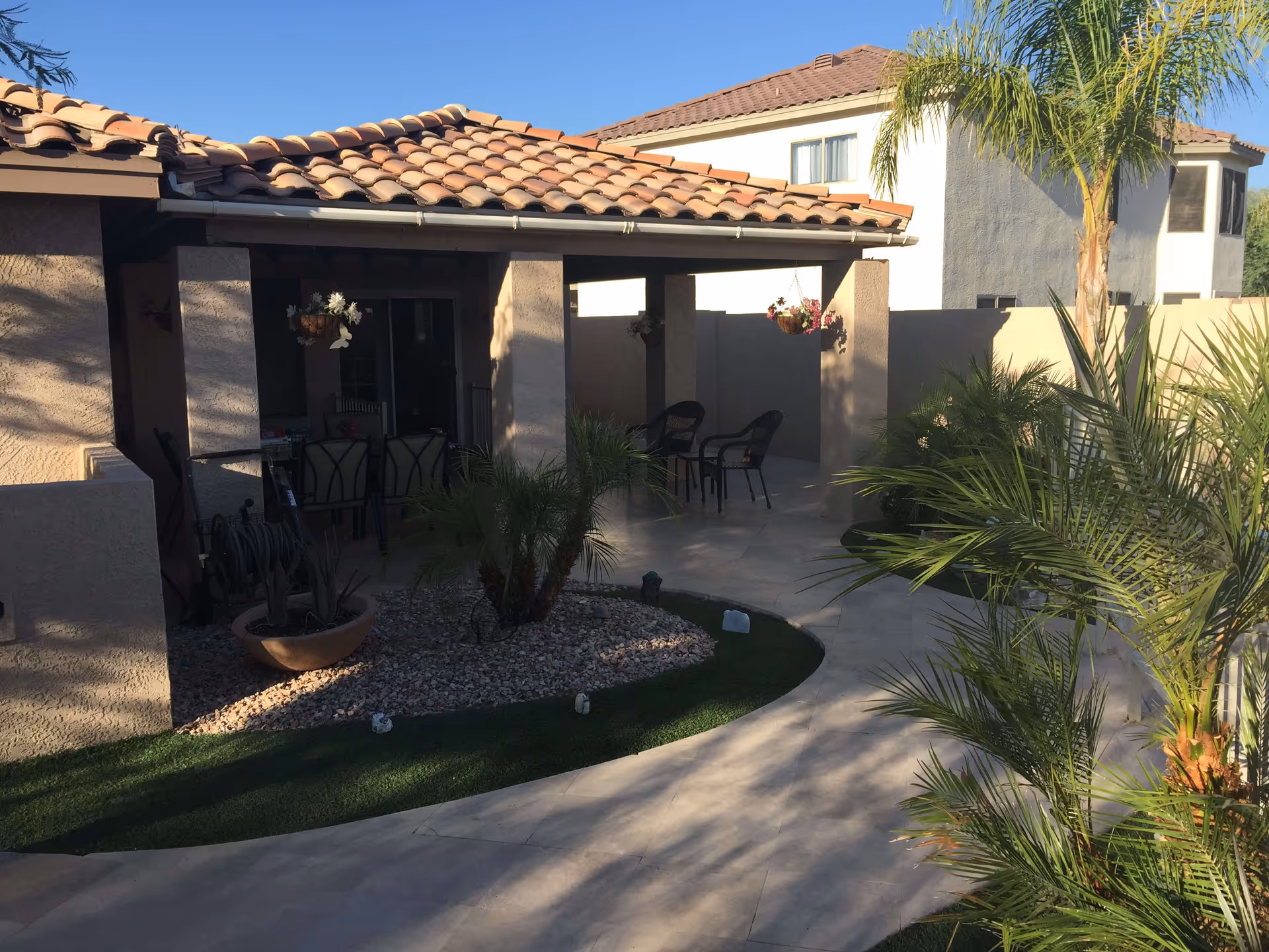Outdoor patio area of a residential building with a tiled roof, beige walls, and a curved walkway surrounded by green plants and palm trees. There are chairs and hanging flower pots under the covered patio.