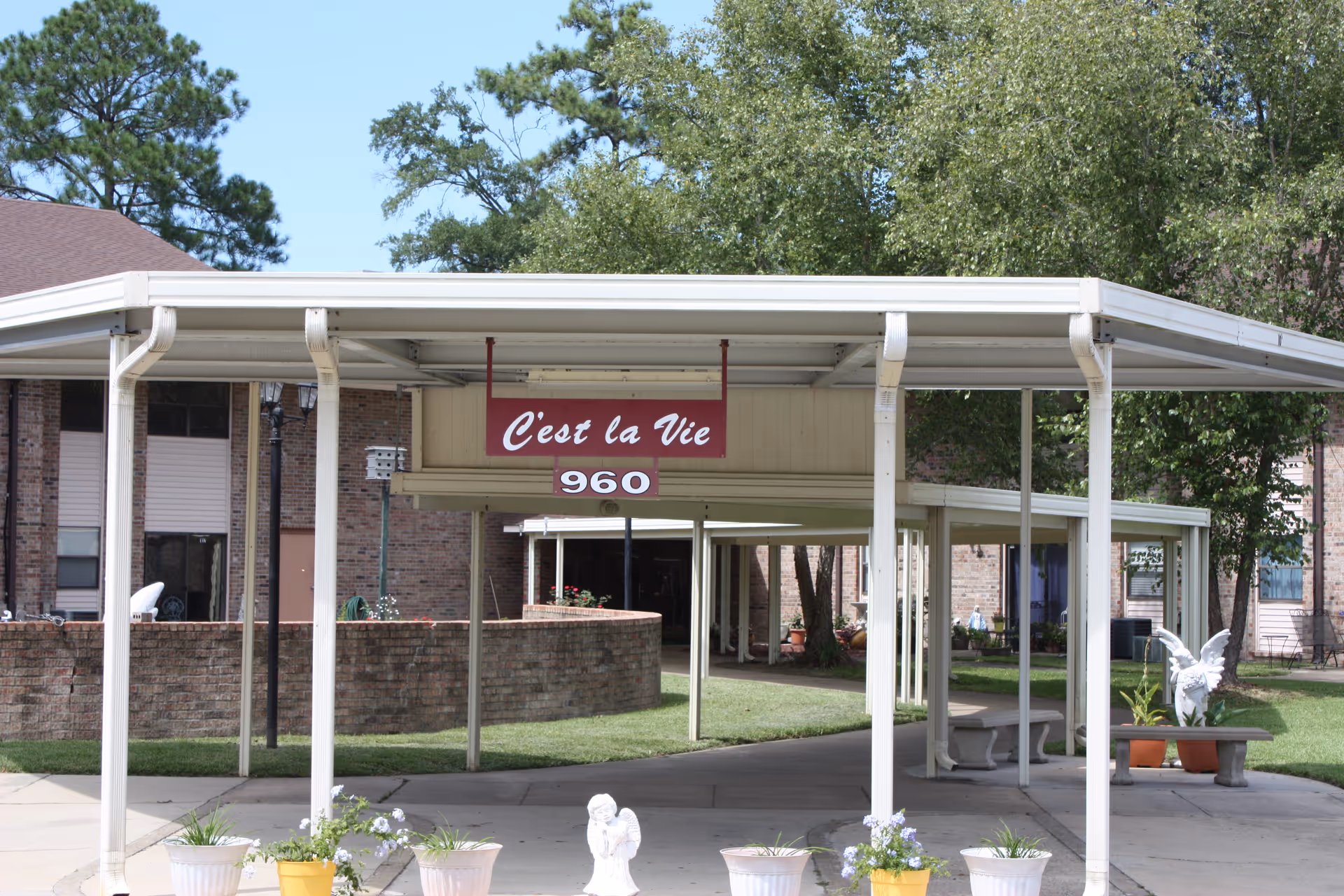 Covered walkway leading to the entrance of a brick building with a sign that reads 'C'est la Vie 960'. The area is landscaped with grass, trees, and several potted plants and small statues, including angel figures.
