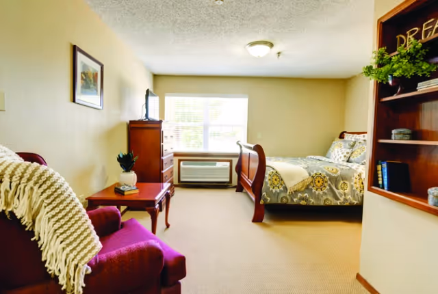 Bright furnished bedroom with a wooden sleigh bed, armchair, dresser, window AC unit and bookshelf.