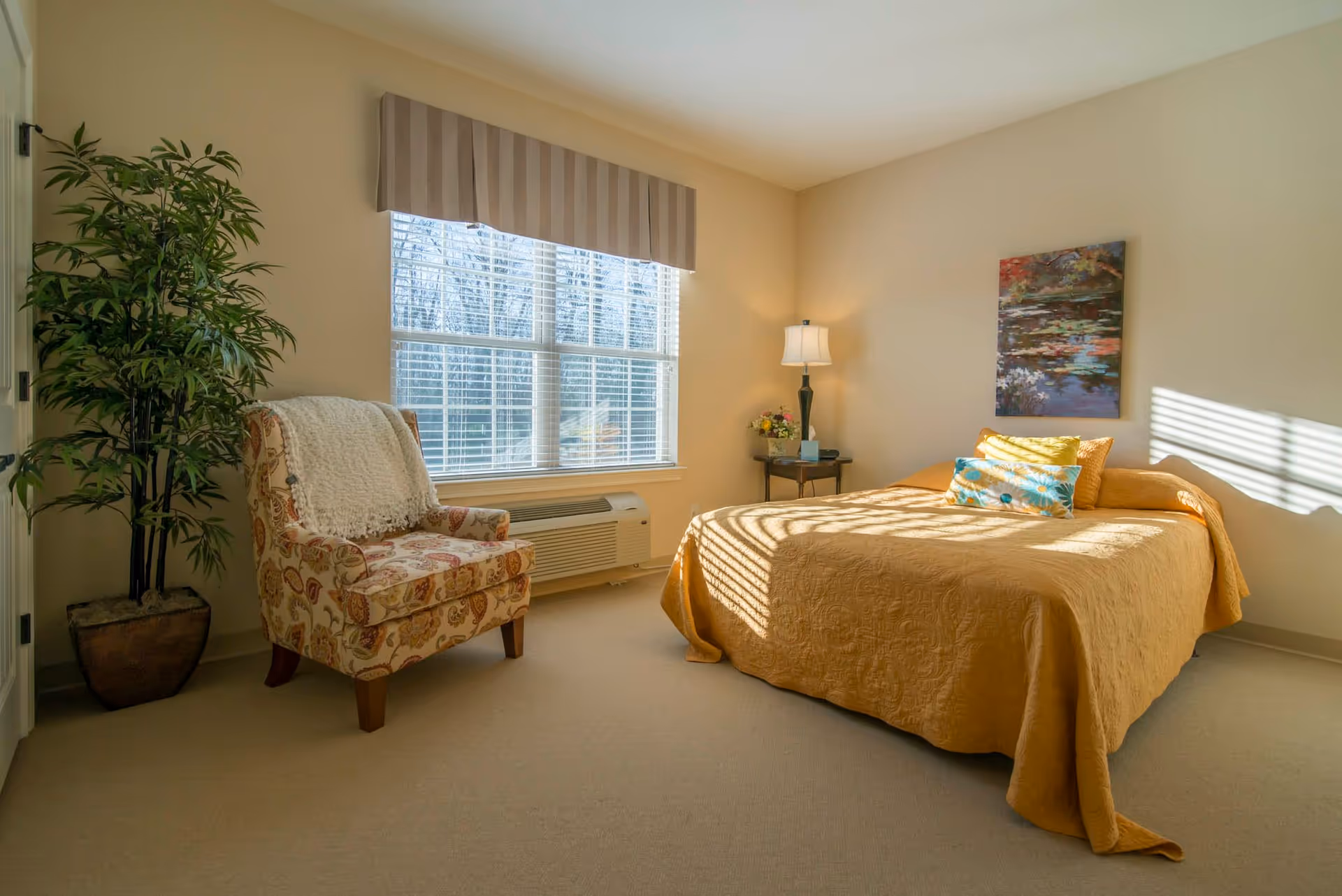 Sunlit bedroom with a bed covered in a golden quilt, a patterned armchair, potted plant, window with blinds, and a bedside table with a lamp.