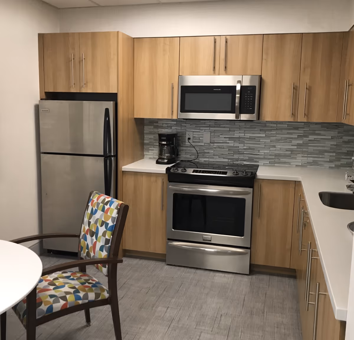 Compact kitchen with wooden cabinets, stainless appliances, and a patterned chair.