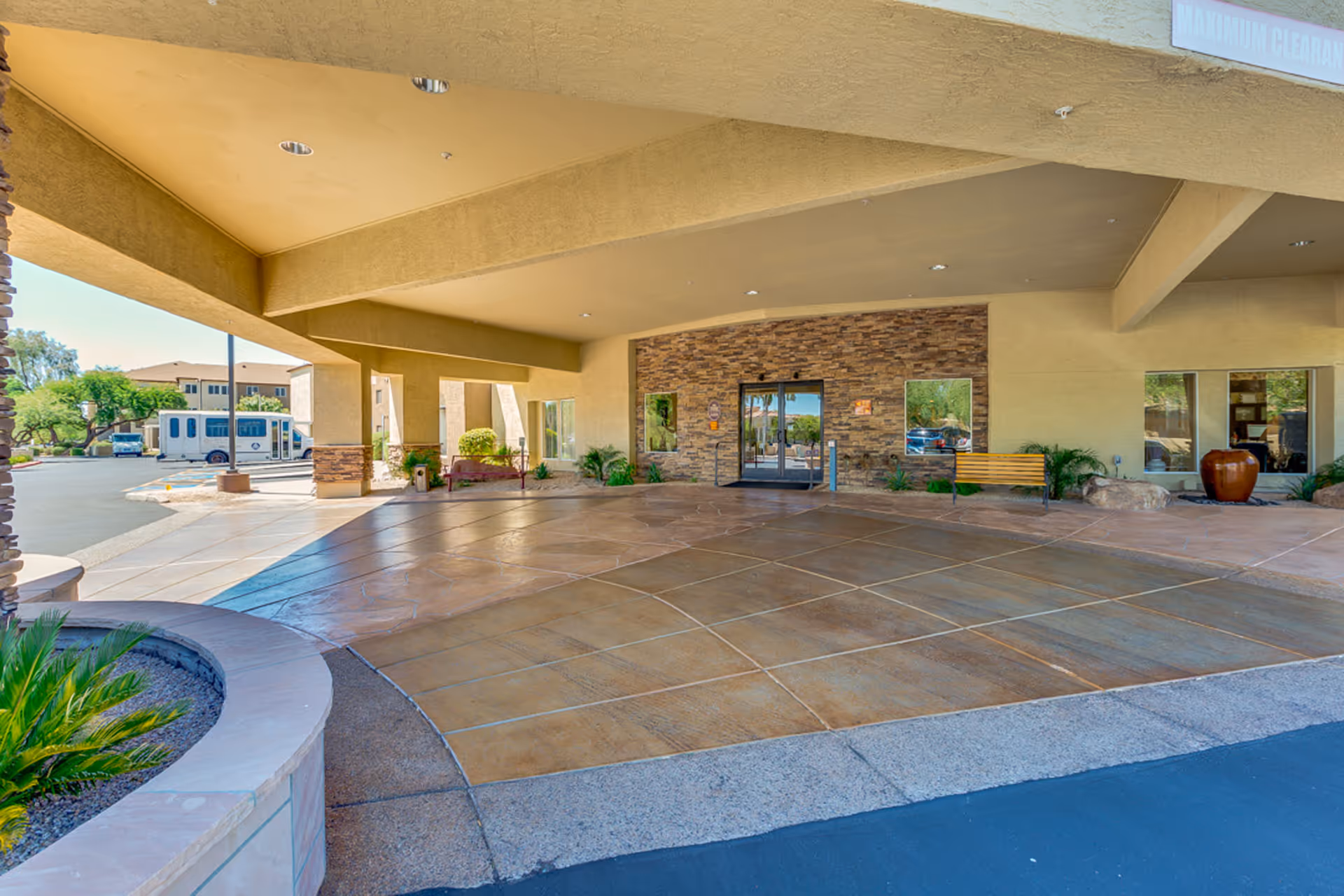 Covered entrance area of Discovery Point Retirement Community with a stone accent wall, glass double doors, a wooden bench, potted plants, and a driveway for vehicle drop-off. A shuttle bus is visible in the background.