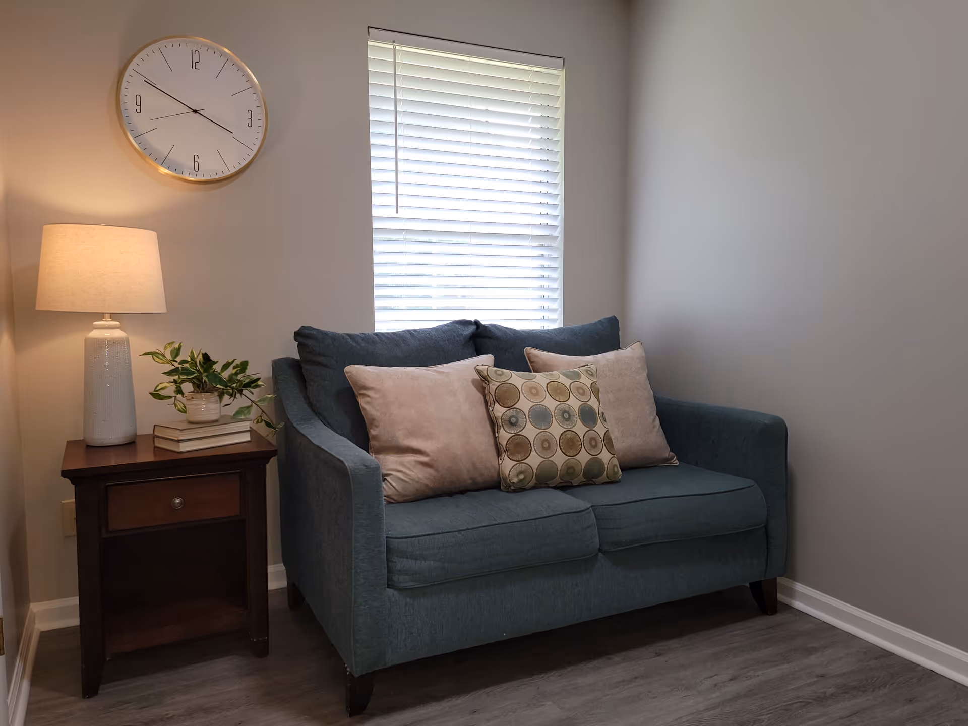 Teal sofa with cushions beside a wooden side table, lamp, clock, and window blinds.