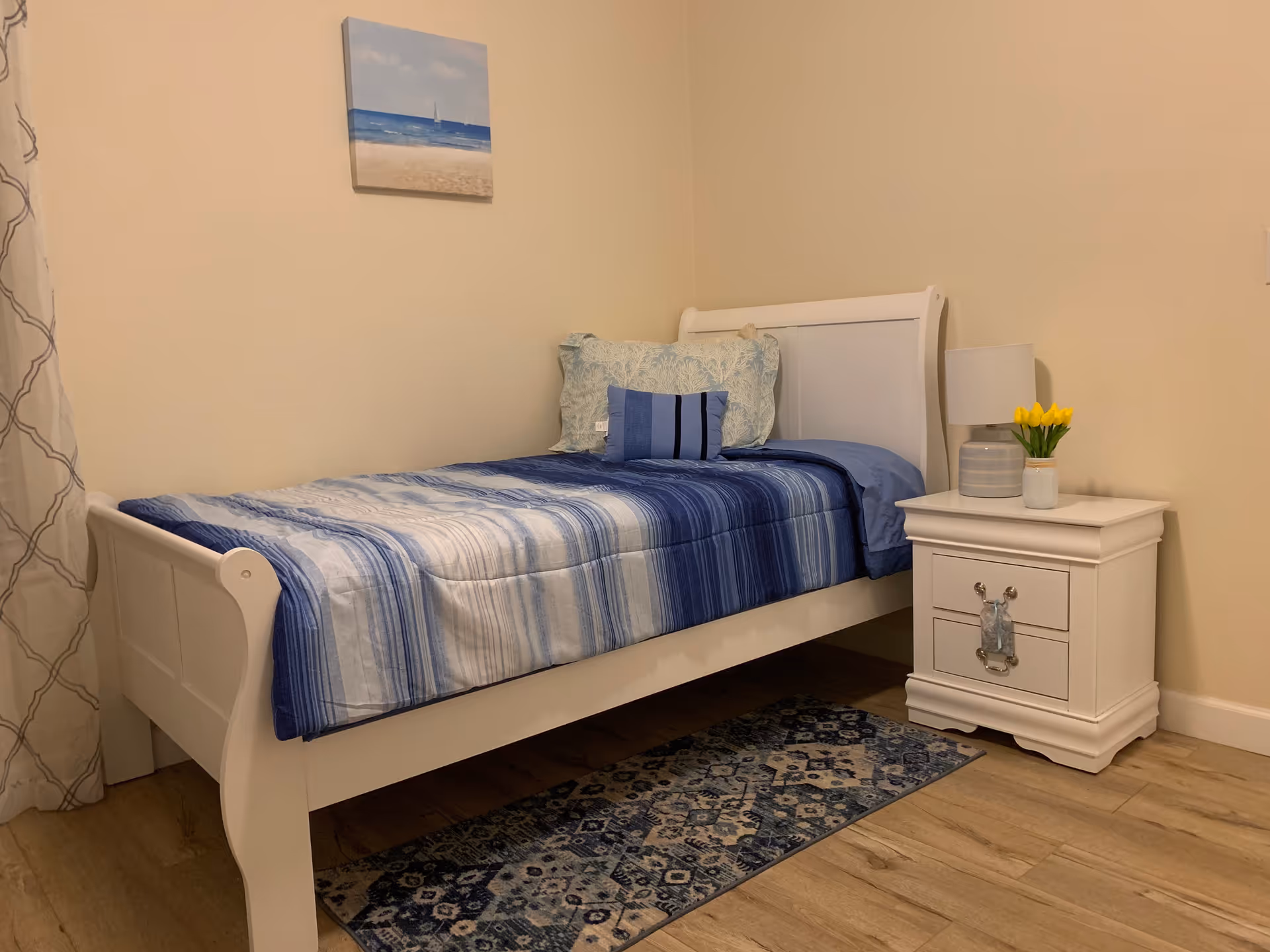 A small bedroom with a white single bed featuring blue and white striped bedding and multiple pillows. Next to the bed is a white nightstand with a lamp and a small vase of yellow tulips. A blue patterned rug is on the wooden floor, and a beach-themed painting hangs on the beige wall above the bed.