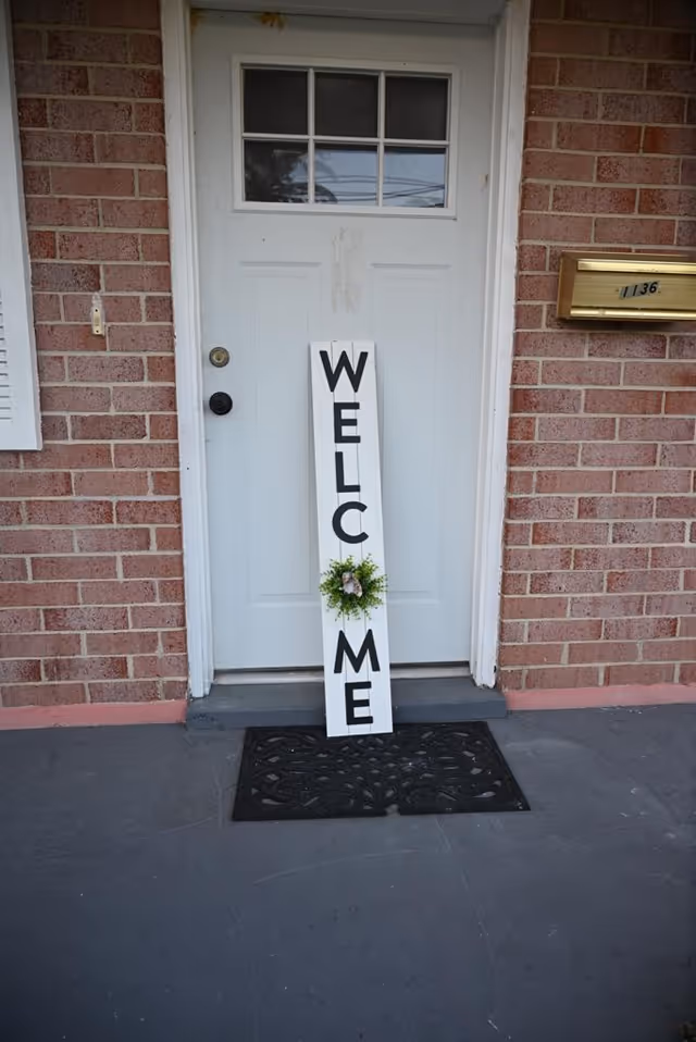 Front door on brick exterior with a tall vertical 'WELCOME' sign and wreath.