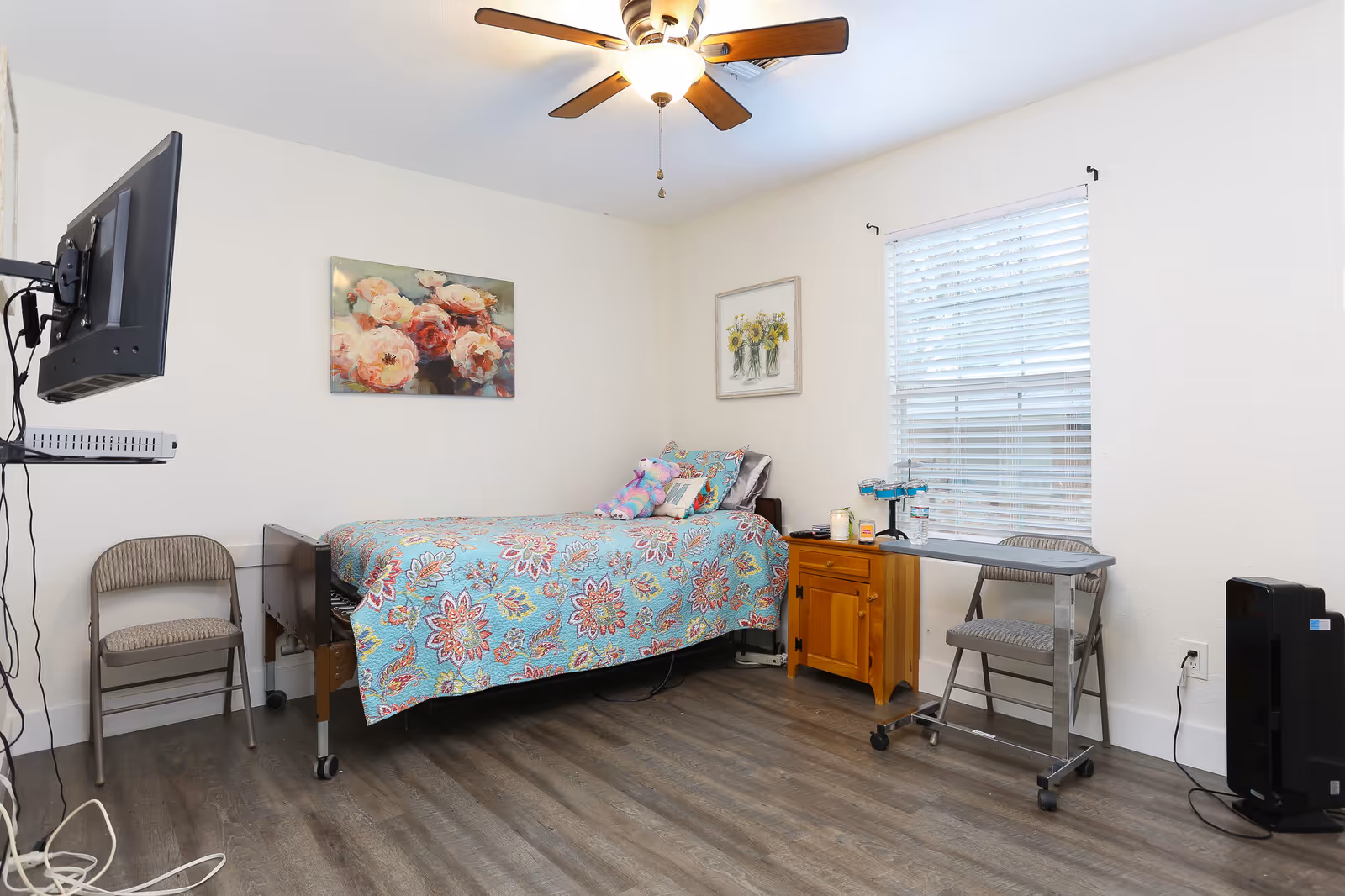 Furnished bedroom in a senior living facility with a bed, desk, and floral decor.