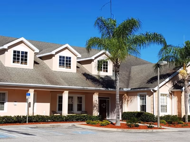 Front exterior of a pale-pink building with dormer windows, palm trees, and a parking area.