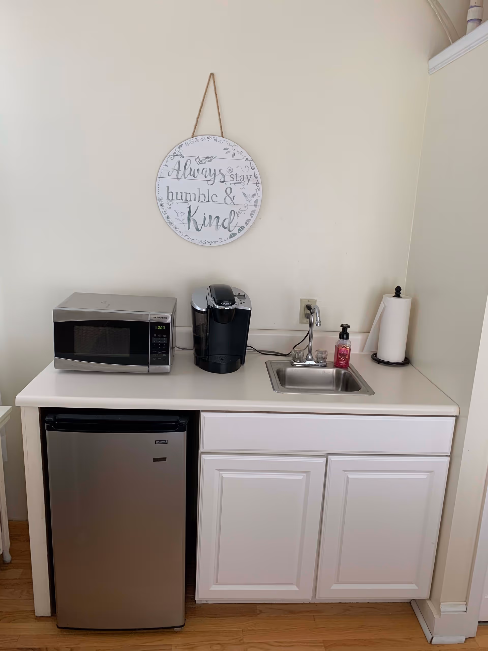 A small kitchenette area with a stainless steel mini fridge, a microwave, a Keurig coffee maker, a small sink with a faucet, a bottle of hand soap, and a paper towel holder. Above the counter, a round decorative sign hangs on the wall that reads 'Always stay humble & kind.' The cabinetry is white, and the floor is wooden.