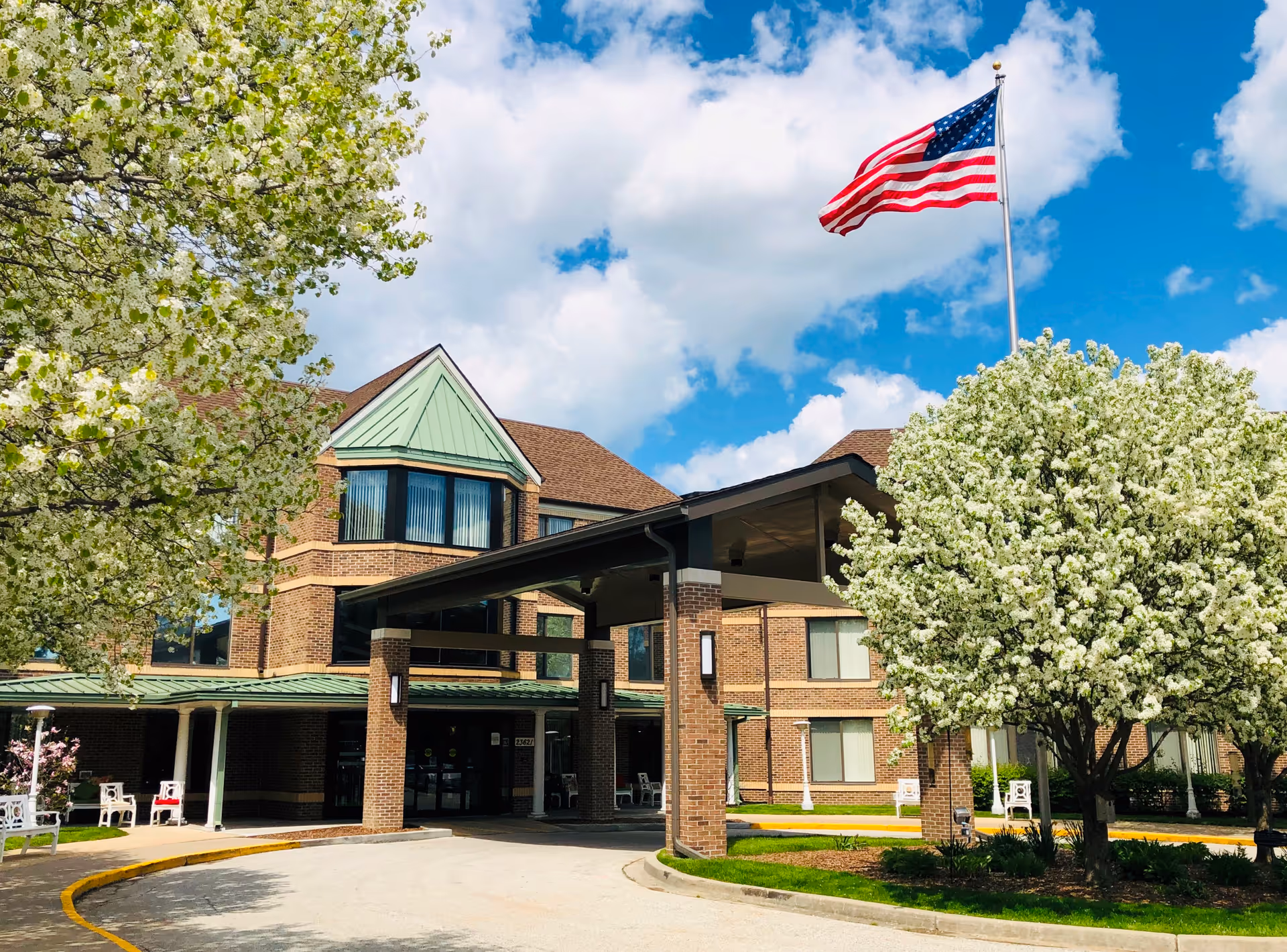 Brick senior living facility with a covered entrance, blooming trees, and a waving American flag