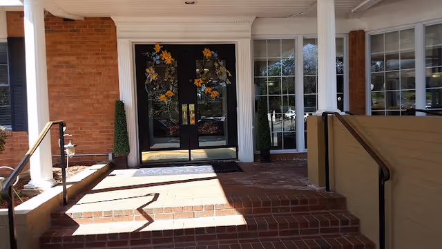 Brick entrance with steps leading to double glass doors decorated with wreaths, flanked by white columns and large windows.
