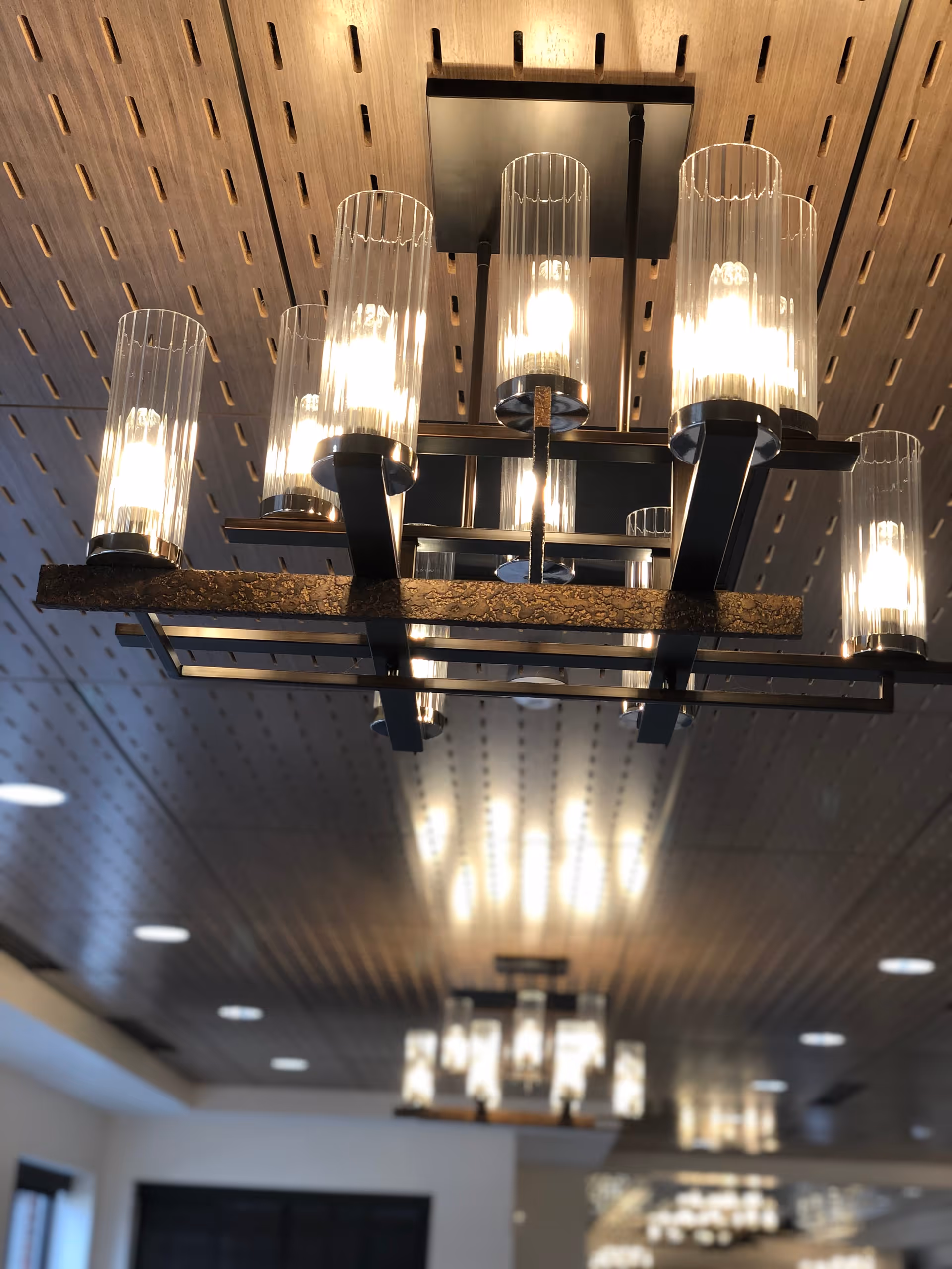 Modern ceiling light fixture with multiple cylindrical glass shades and lit bulbs, mounted on a wooden slatted ceiling in an interior space.