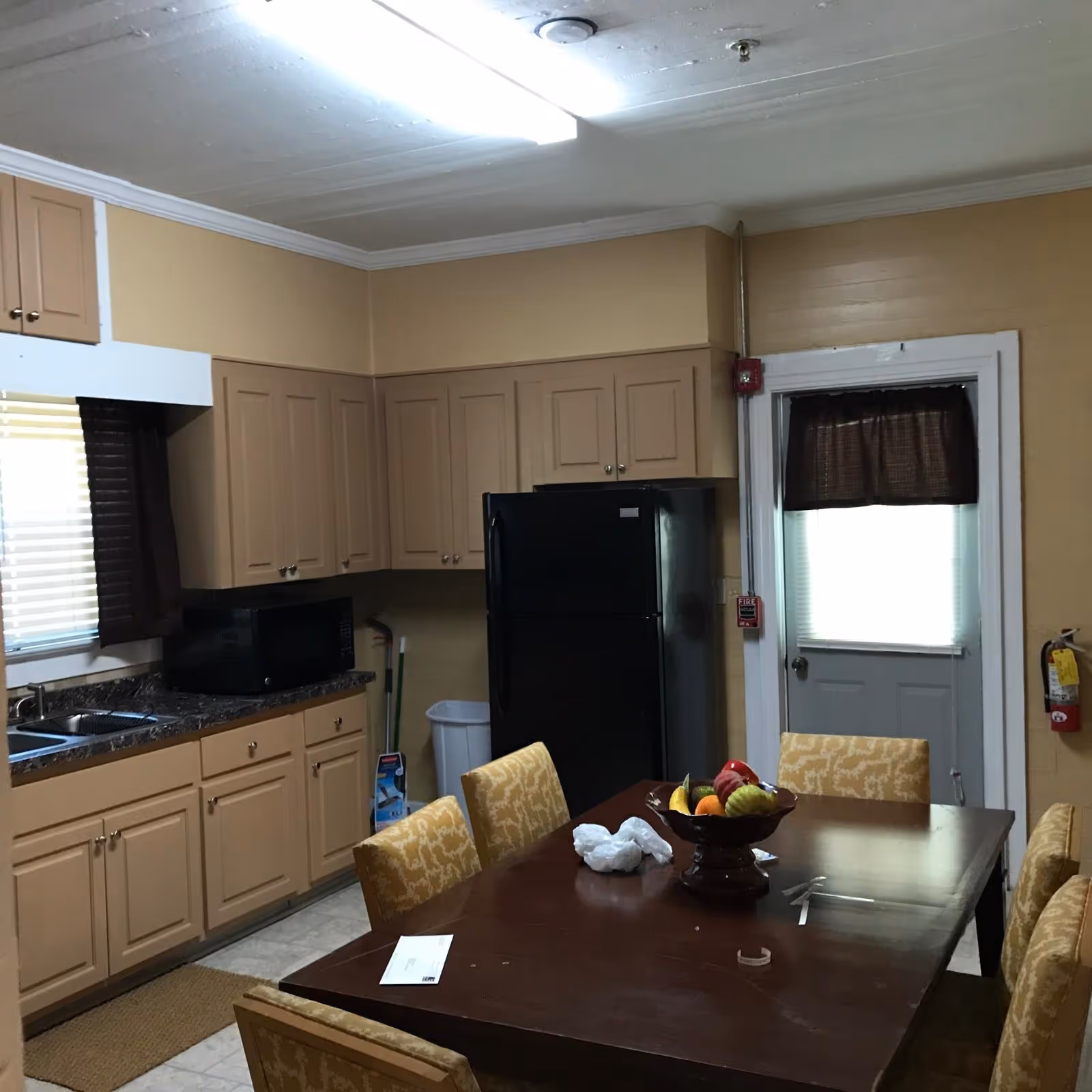 Kitchen and dining area with beige cabinets, a dark dining table, fruit bowl, chairs, and fridge.