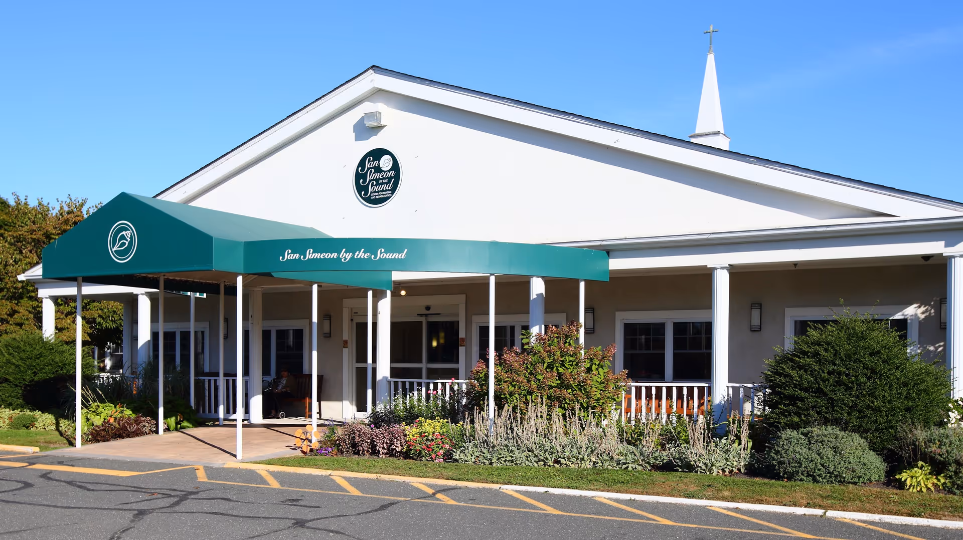 White senior living facility exterior with a green canopy entrance and landscaped front garden.
