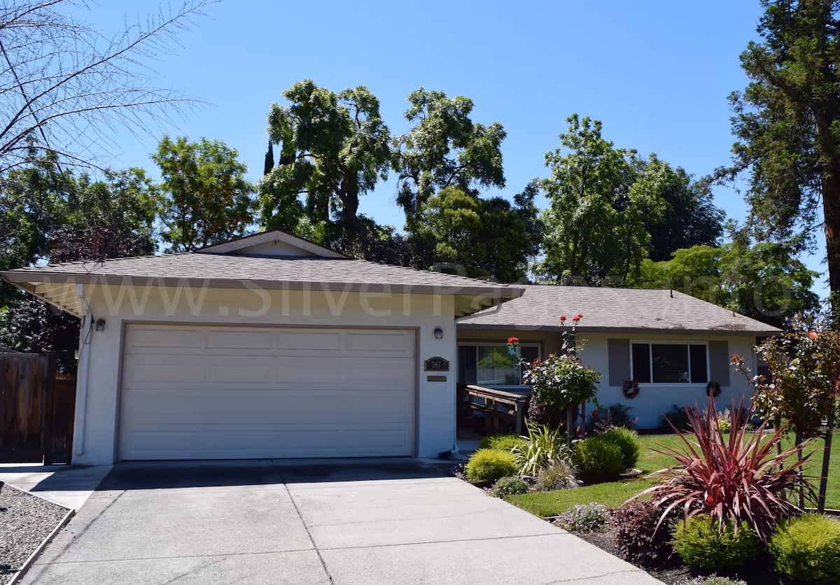 Single-story house with a two-car garage, a concrete driveway, and a well-maintained front yard with various plants and shrubs under a clear blue sky.