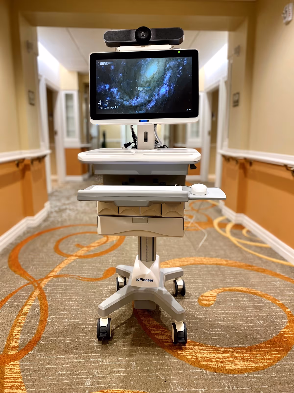 Telemedicine cart with monitor and camera stands in a beige hallway of a senior living facility.