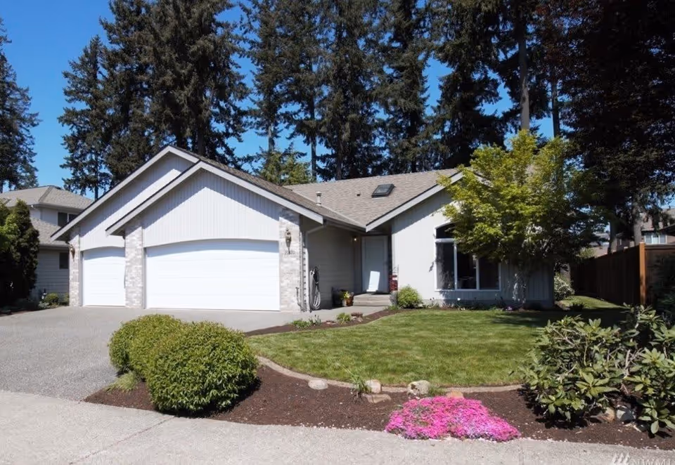Single-story house with a three-car garage, manicured front lawn, shrubs and trees under a clear blue sky.