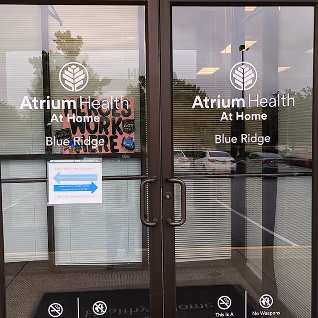Glass entrance doors display Atrium Health At Home Blue Ridge signage and reflection of parking lot.