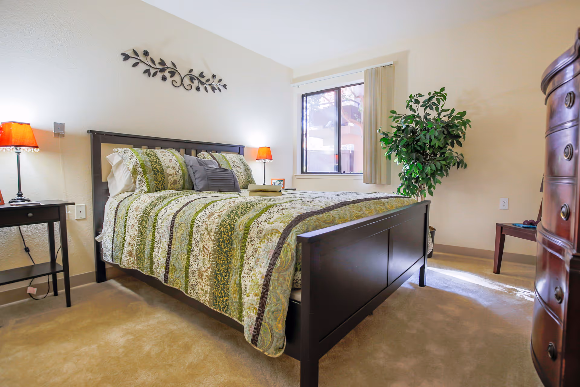A dark wood bed with green quilt, matching nightstands, lamp, and plant in a bright bedroom.