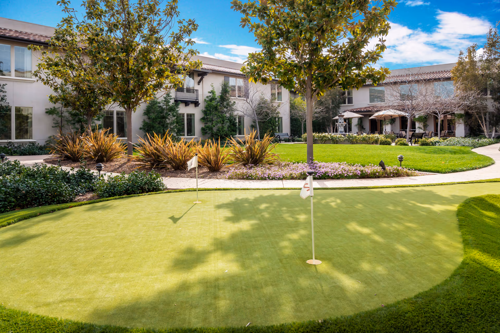 A well-maintained outdoor putting green with two small flags, surrounded by landscaped bushes, trees, and a walking path. In the background, there is a two-story building with large windows and a patio area with tables and umbrellas under a blue sky with some clouds.