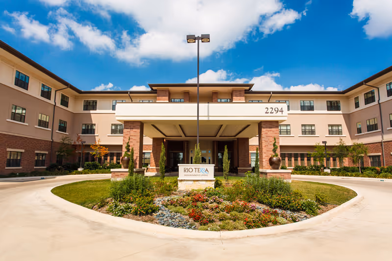 Front entrance of the three-story Rio Terra Senior Living building with a covered porte-cochère, circular driveway and landscaped central island.
