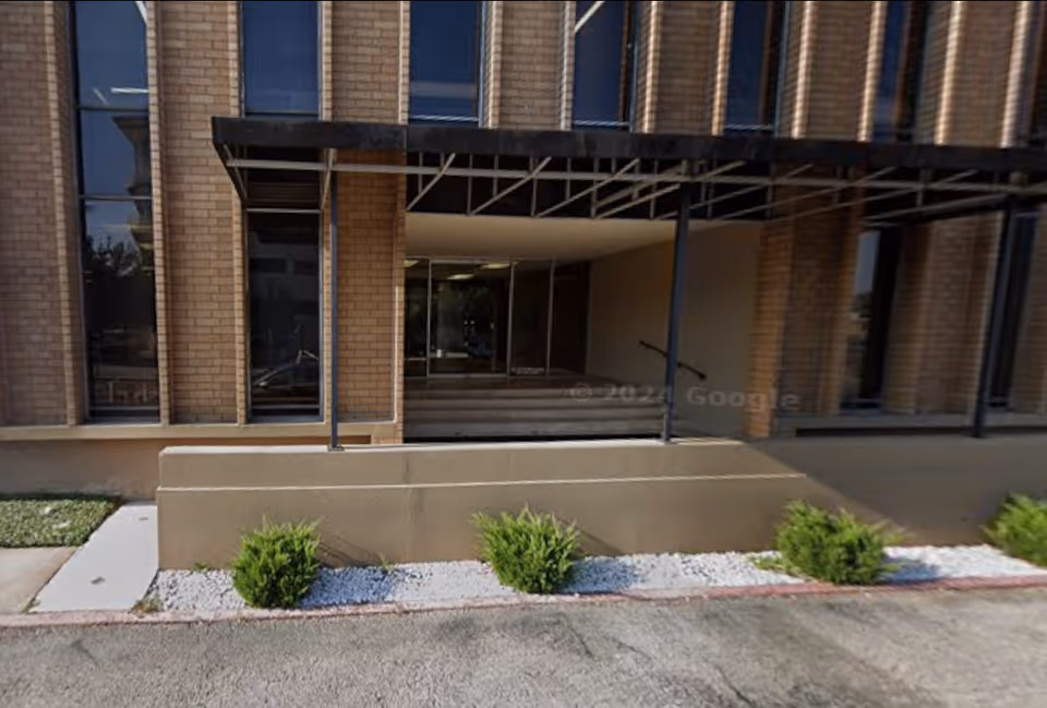 Exterior brick entrance with glass doors, ramp, and canopy; small shrubs along the walkway.