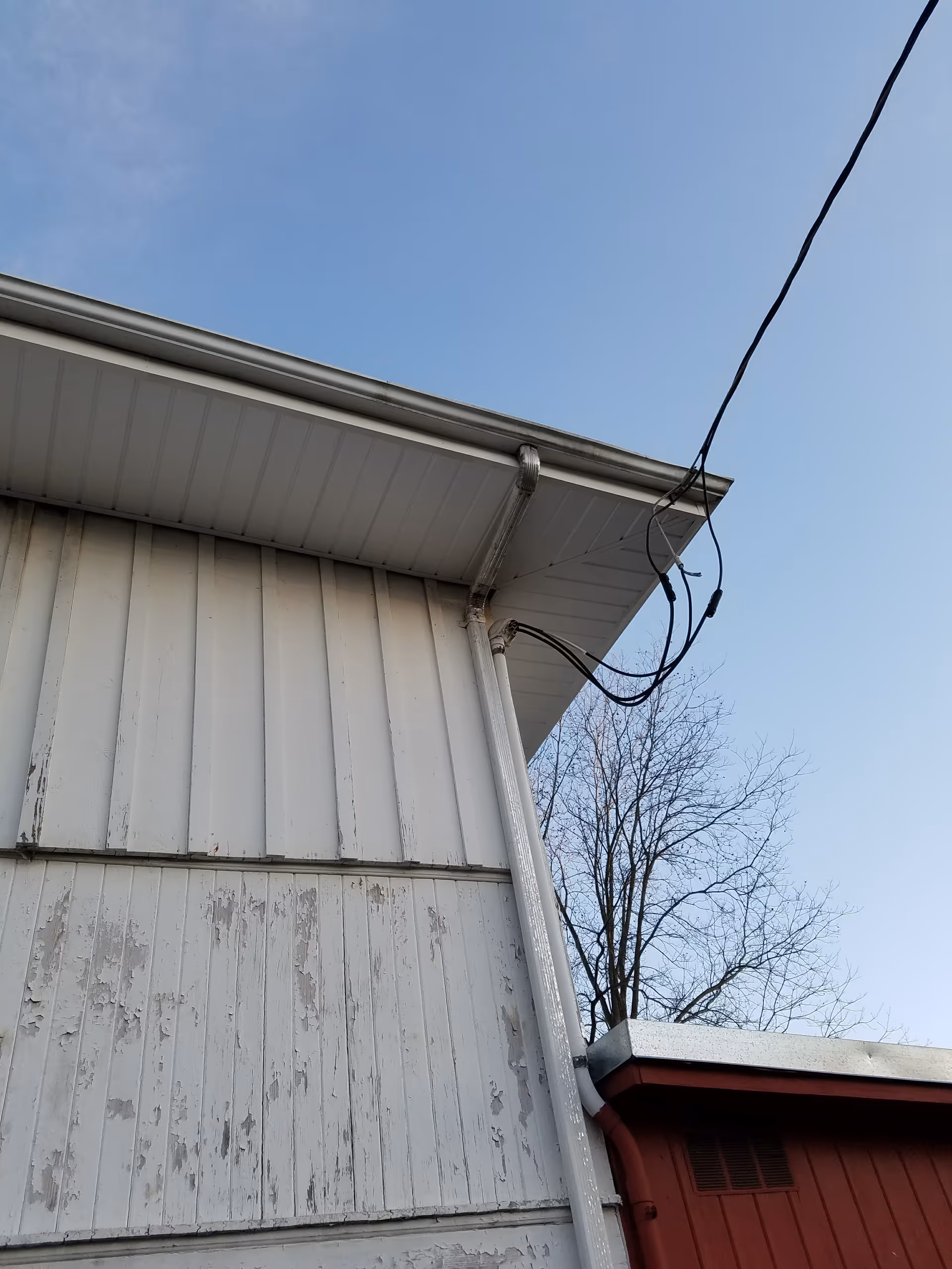 View of peeling white siding, gutter, and hanging wires on a building exterior against blue sky.