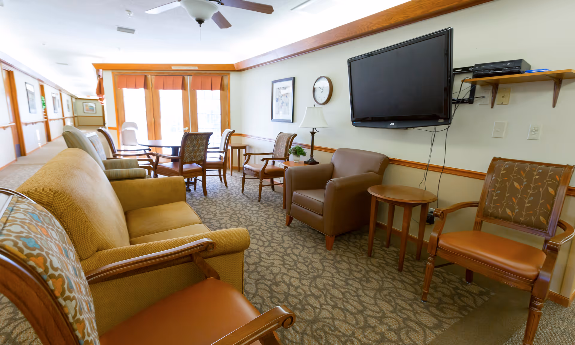 Senior living facility lounge with sofa, chairs, tables, and wall-mounted TV.
