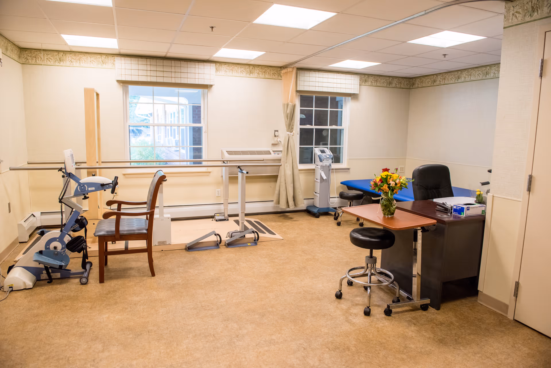 A physical therapy room with exercise equipment including a stationary bike, parallel bars for walking practice, a wooden chair, a blue therapy table, a desk with a black office chair, a small rolling stool, and a vase of flowers on a small table. The room has beige walls, two windows with blinds, and a beige floor.