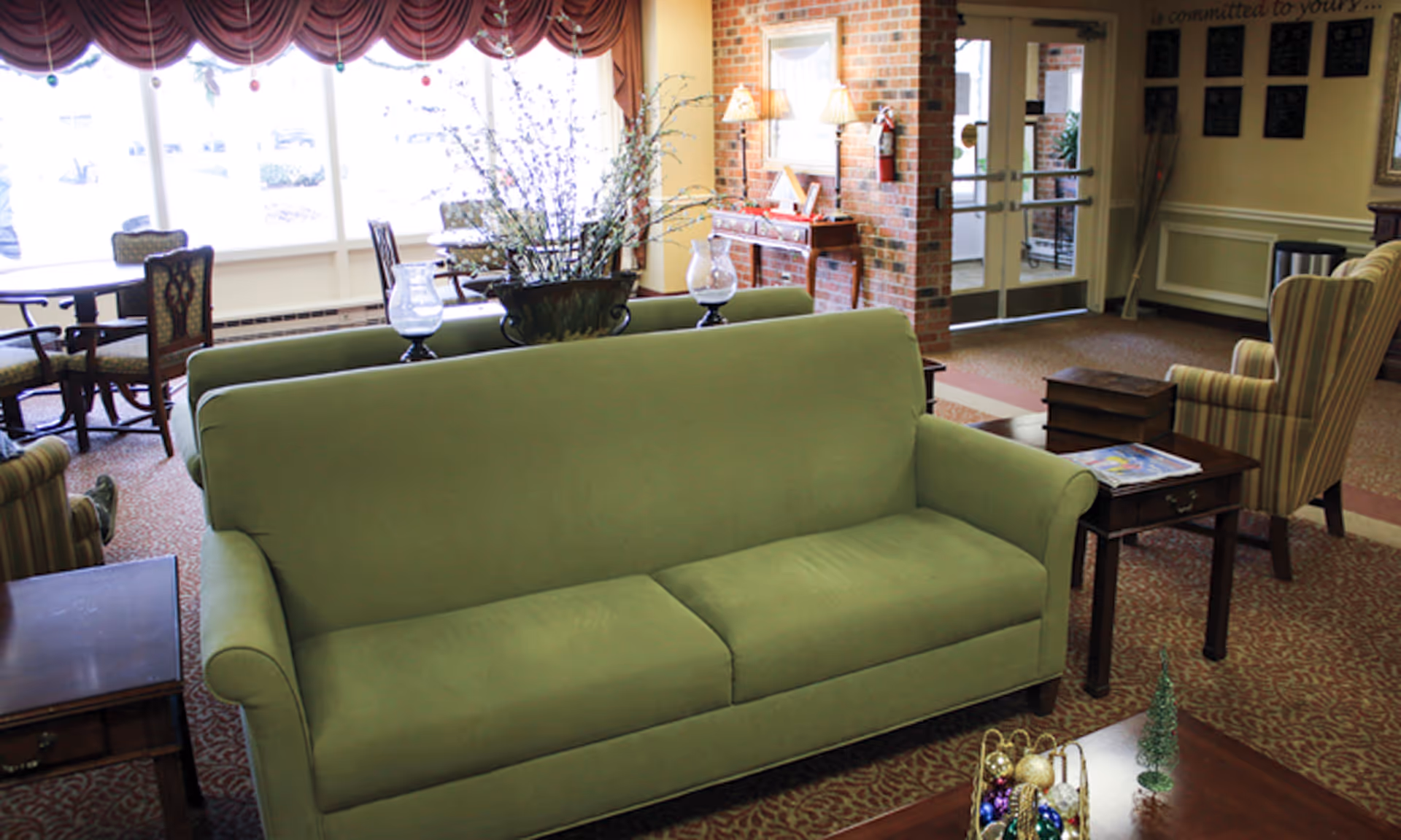 Green sofa anchors a bright senior living lounge with chairs, tables, and large windows.