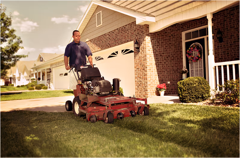 A man mows a well-kept lawn in front of a brick house with a wreath.