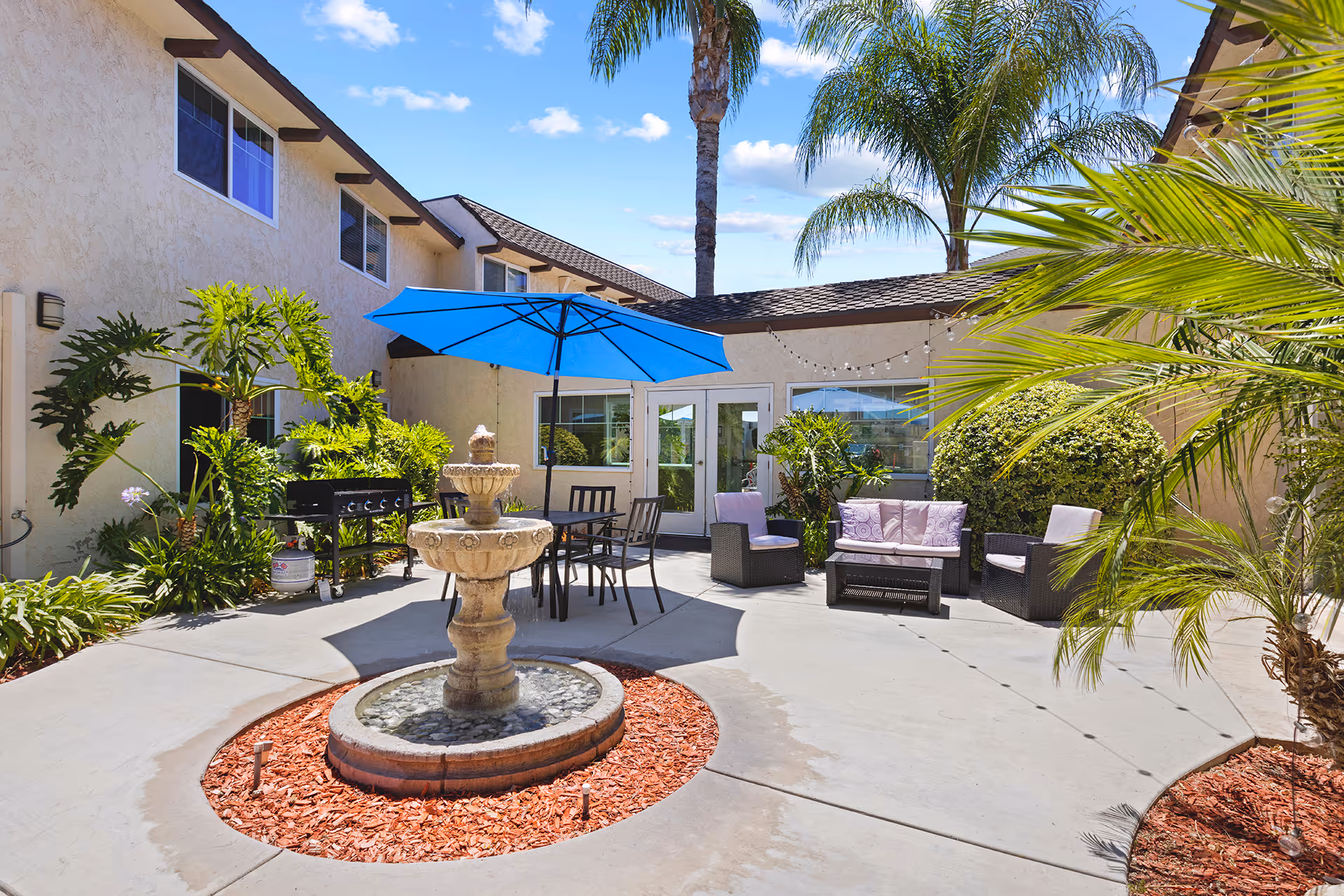 Outdoor courtyard area at a senior living facility with a central stone water fountain surrounded by red mulch. There is a patio table with a blue umbrella and chairs, a seating area with cushioned chairs and a loveseat, various green plants, palm trees, and a beige building with windows and doors in the background under a blue sky with some clouds.