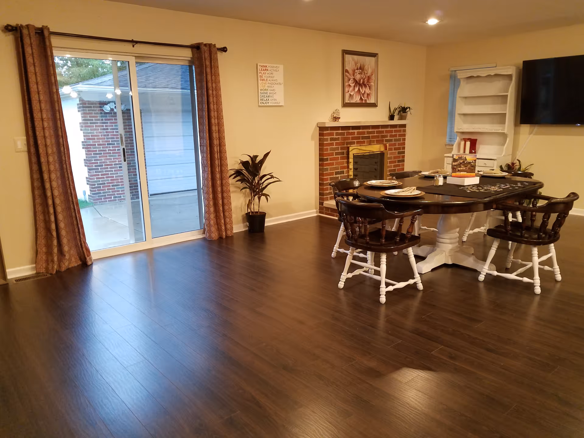 Interior room with dark wood flooring, a sliding glass door with brown curtains leading outside, a brick fireplace with a floral painting above it, a round dining table with four chairs set with plates and napkins, a white cabinet, and a wall-mounted TV.