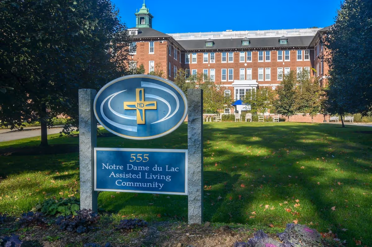 Outdoor view of Notre Dame du Lac Assisted Living Community building with a large sign in the foreground displaying the facility name and address, surrounded by green grass, trees, and outdoor seating.