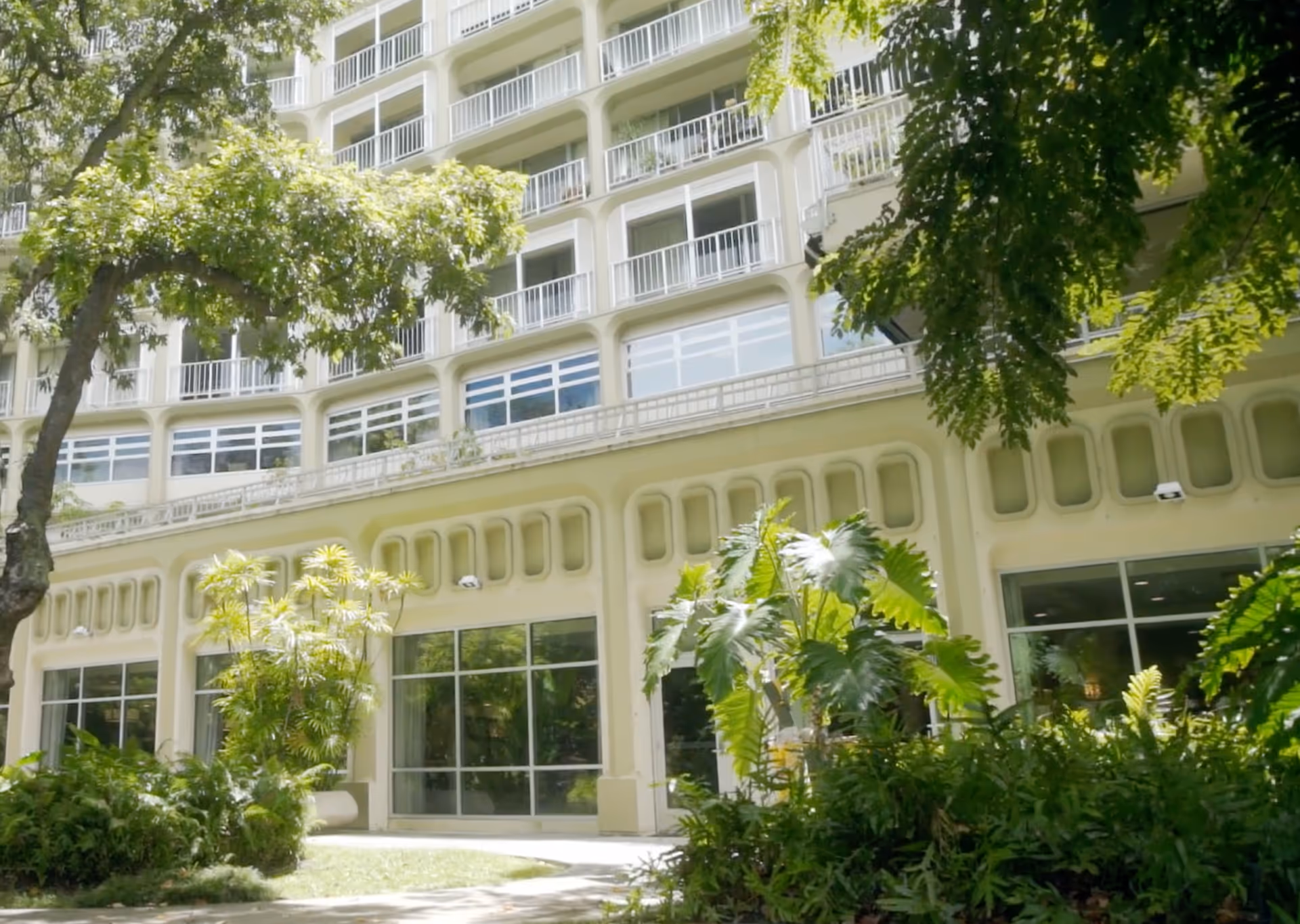 Exterior front of a multi-story retirement residence building with balconies, large windows, and landscaped trees and plants in front.