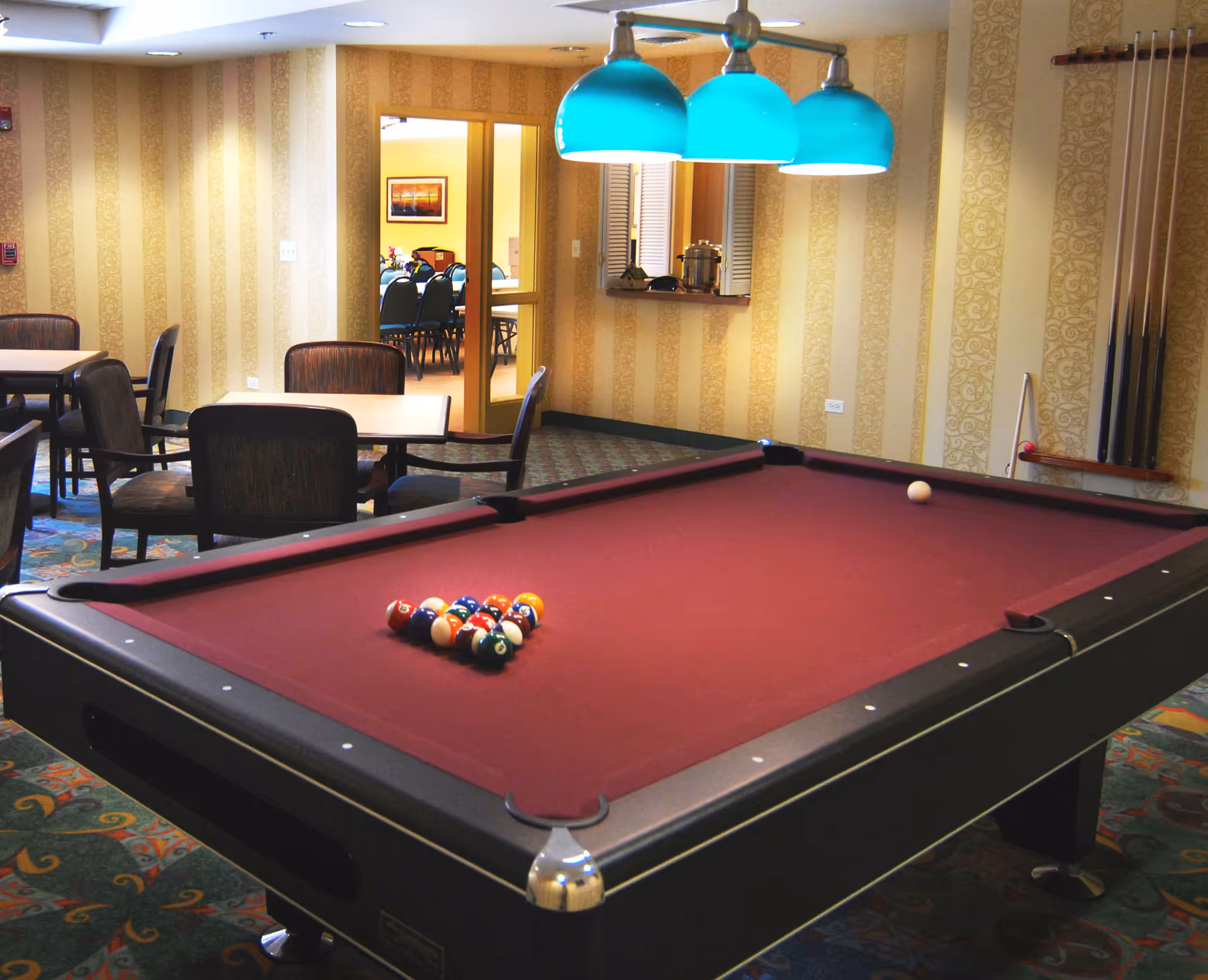 A billiards/recreation room with a burgundy pool table, teal hanging lights, cue rack, and tables and chairs in the background.
