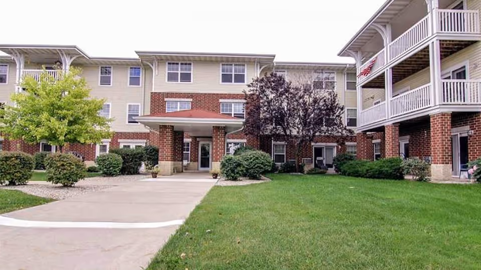 Exterior view of Bell Tower Place Senior Apartments showing a three-story building with beige siding and red brick accents. The building features multiple windows, a covered entrance with a red roof supported by brick columns, balconies on the right side, and landscaped greenery including trees and bushes surrounding a concrete walkway and green lawn.
