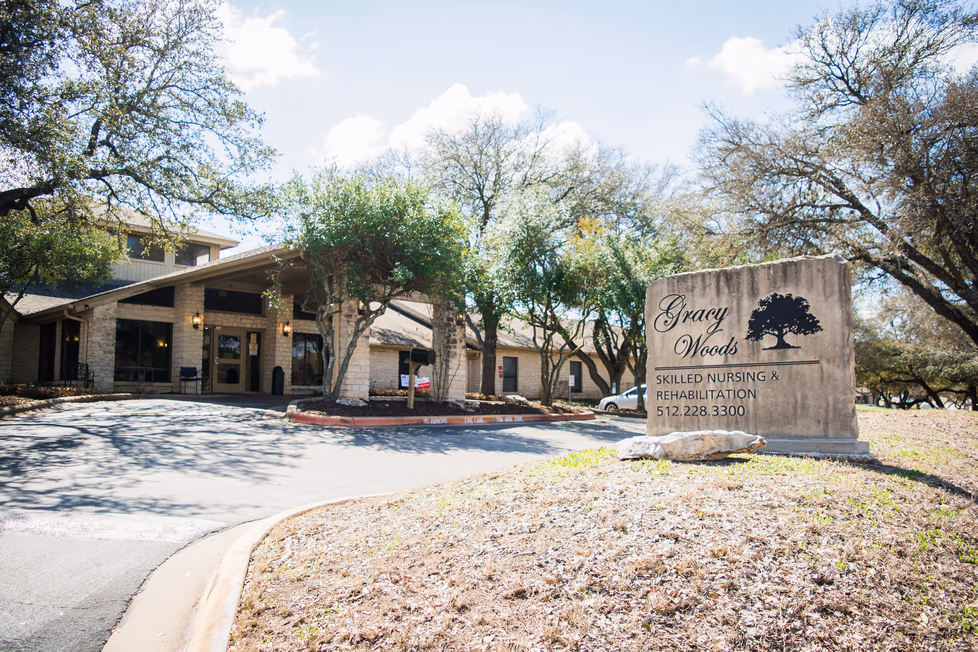 Exterior view of Gracy Woods I Nursing Center showing the entrance of the building with stone walls and a covered driveway. There are trees around the building and a large stone sign in the foreground displaying the facility name, services, and phone number.