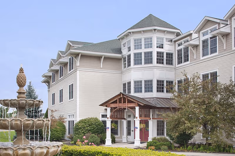 Exterior view of a multi-story senior living facility with beige siding and white trim. The building features a covered entrance with wooden beams and columns, multiple windows, and a green roof. In the foreground, there is a tiered stone fountain with water flowing and landscaped greenery including bushes and trees.